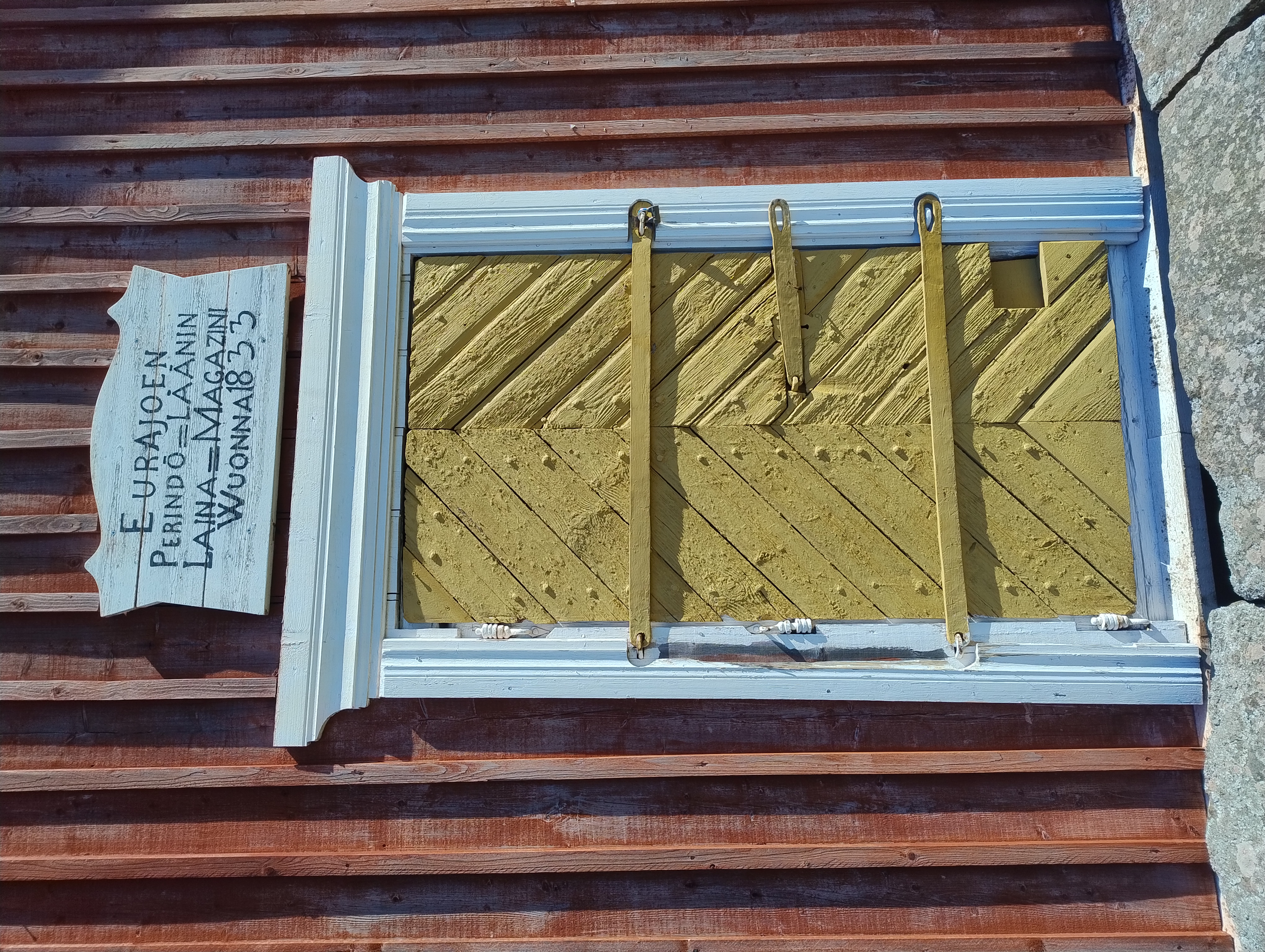 Punainen rakennus, jossa keltainen ovi. Oven yläpuolella kyltti, jossa teksti Eurajoen Perindö-Läänin Laina-Magazini Wuonna 1833