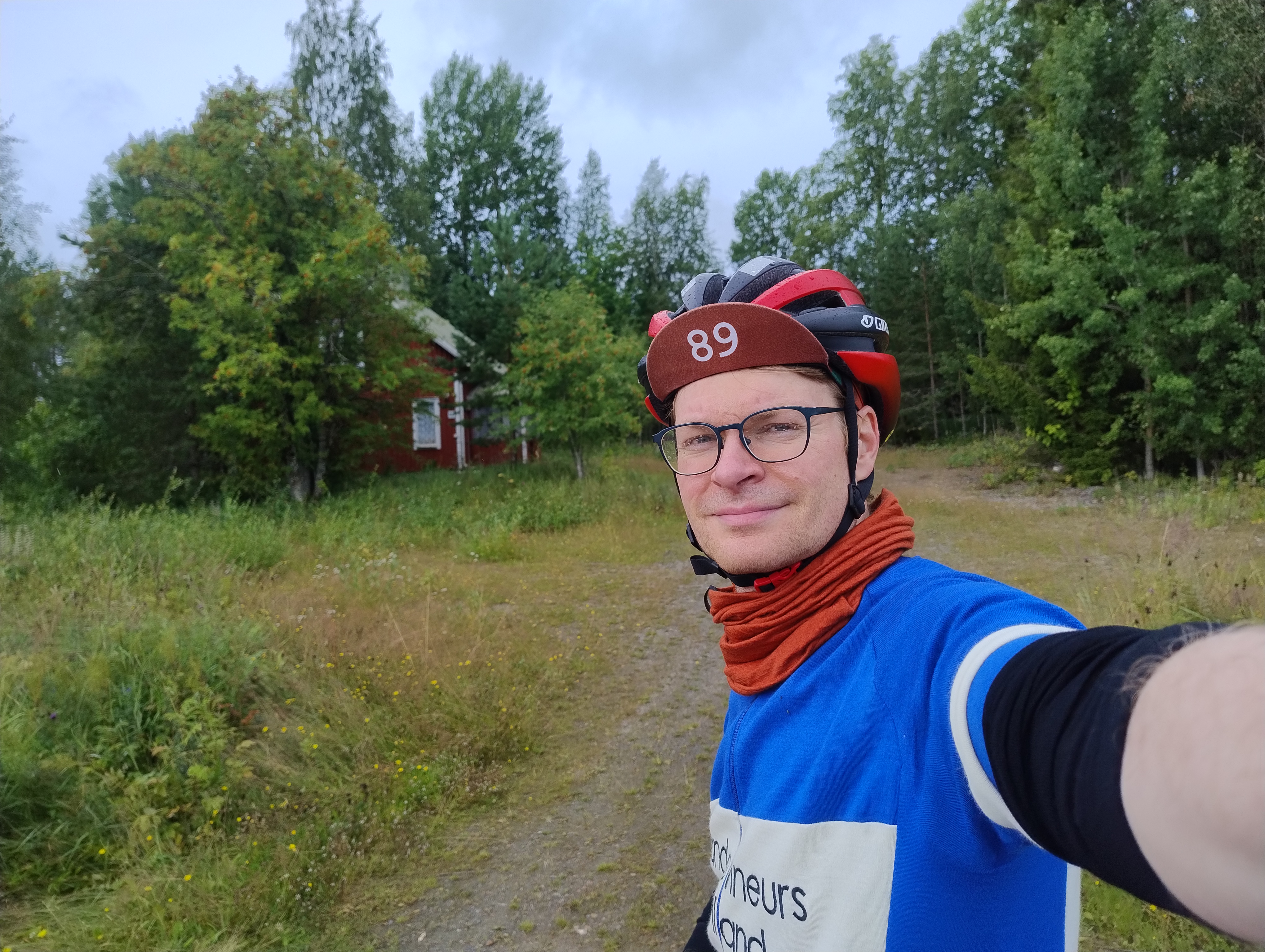 Olli seisoo punaisen talon ja nurmettuneen pihan edustalla. Silmälaseissa on vettä.