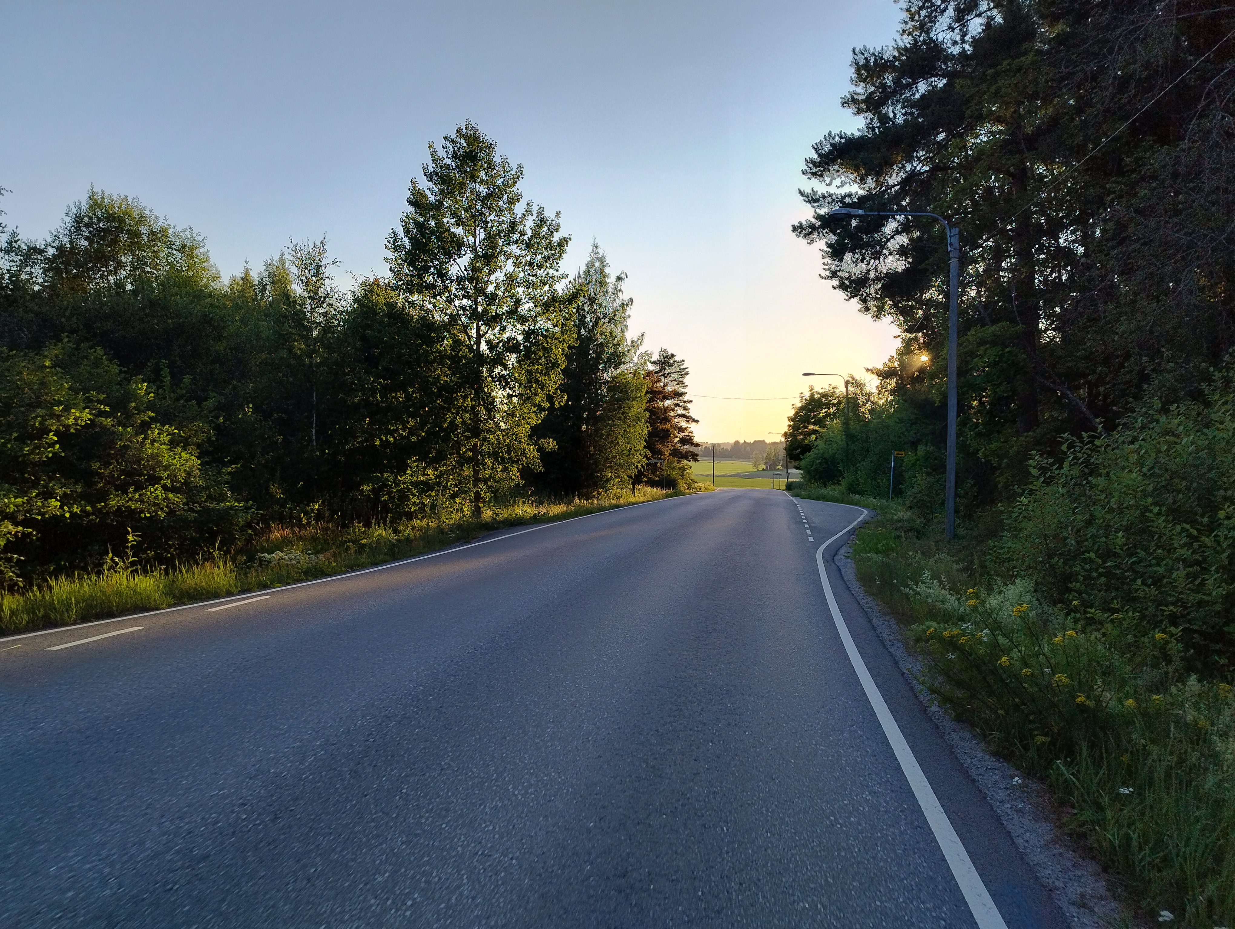 Auringonlasku alaspäin viettävällä asfalttitiellä, edellä aukeaa peltomaisema