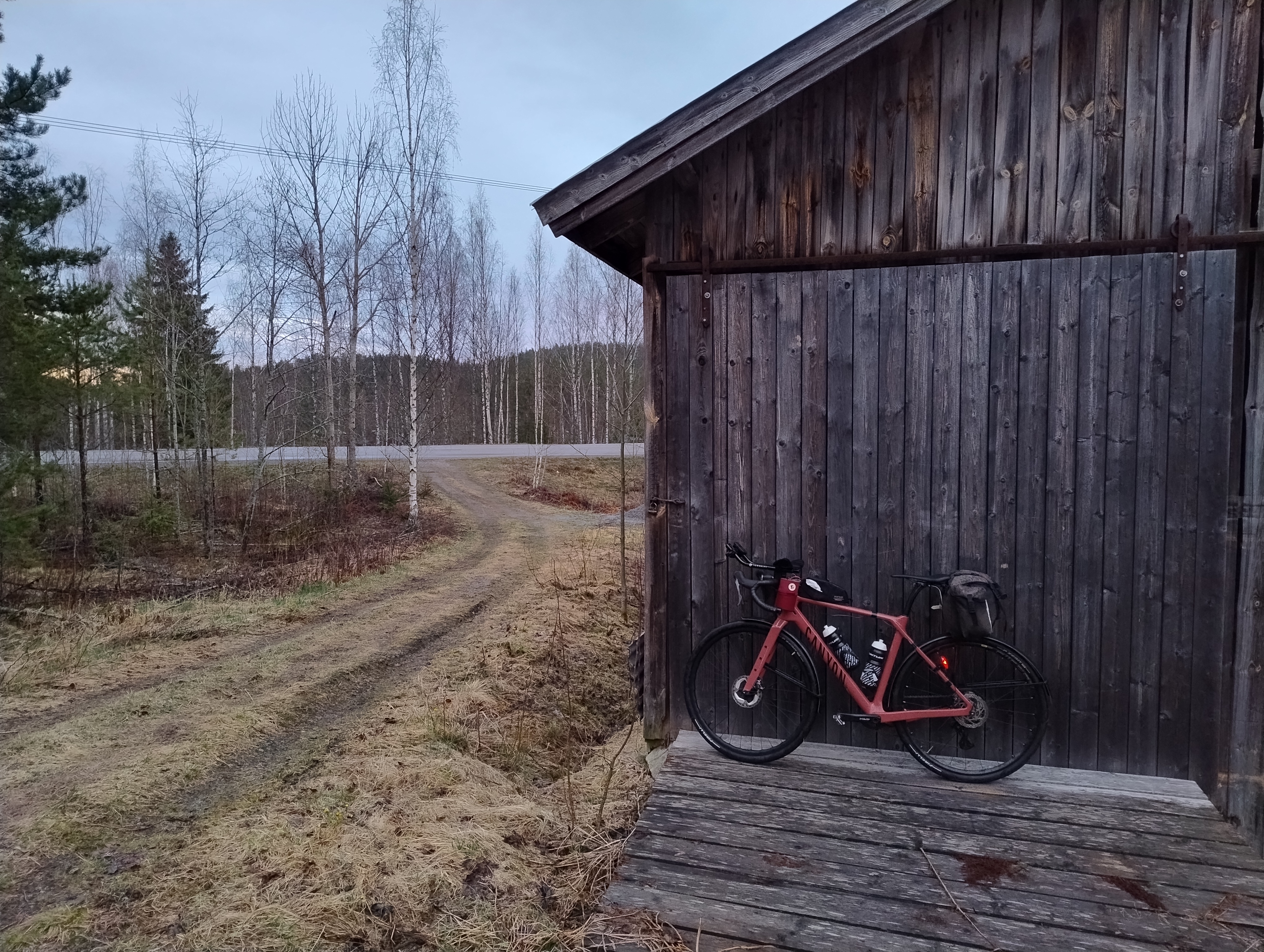 Polkupyörä nojaa harmaan ladon seinään, taustalla asfalttitie