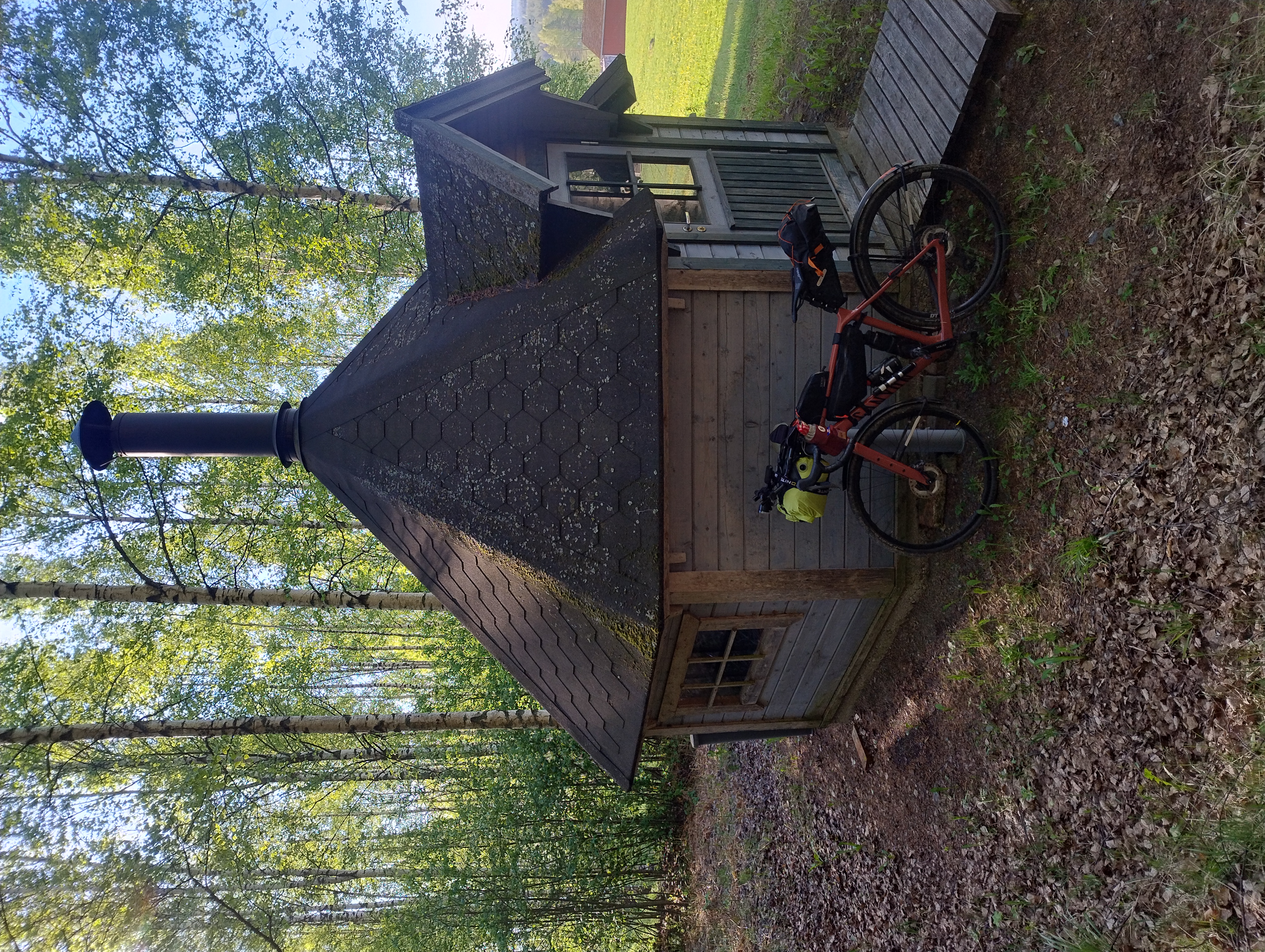 Pyörä nojaa harmaan kodan seinään, taustalla punainen maatalousrakennus.