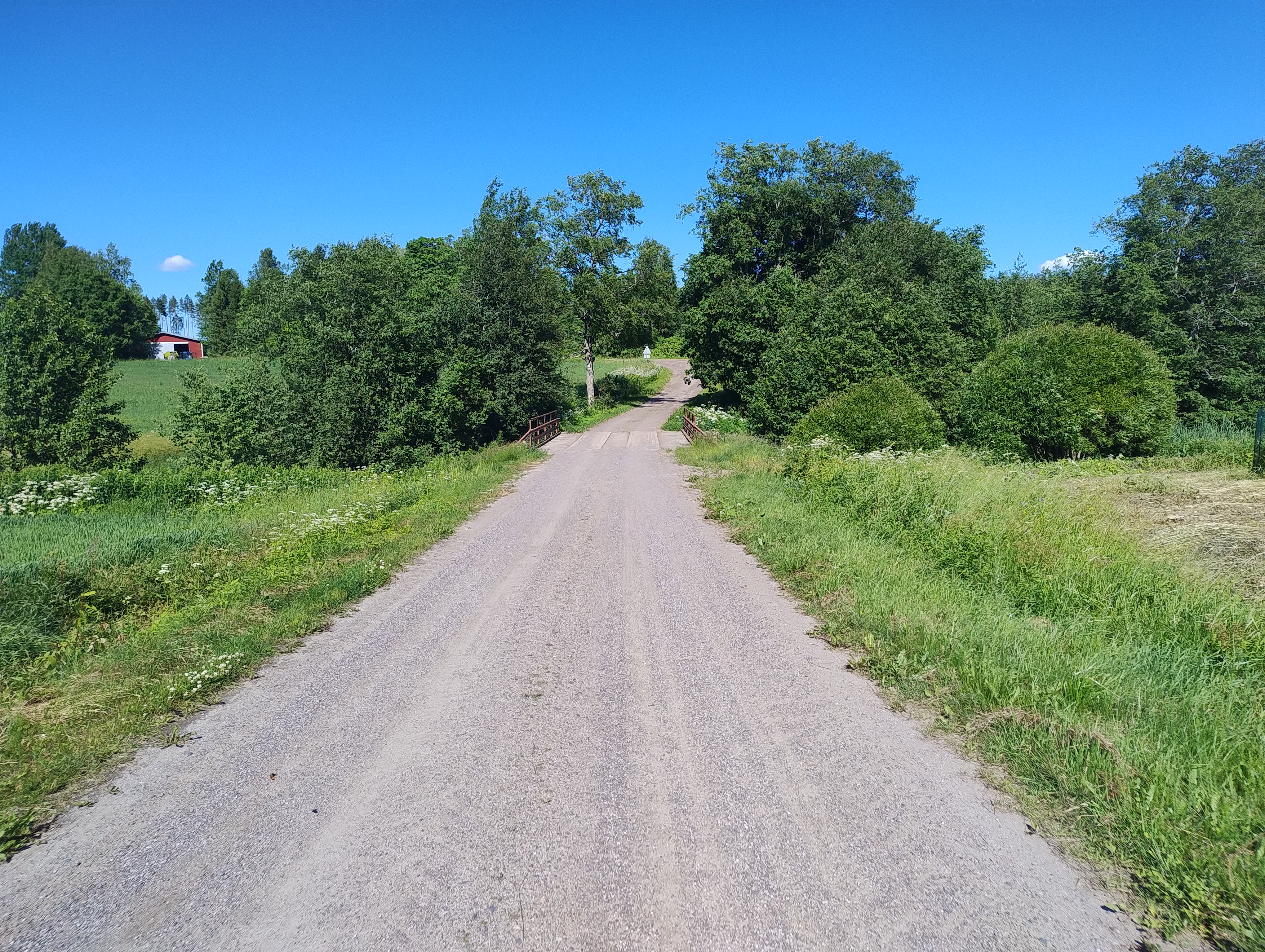 Pieni silta hiekkatiellä auringossa, vieressä peltoja ja matalia puita.