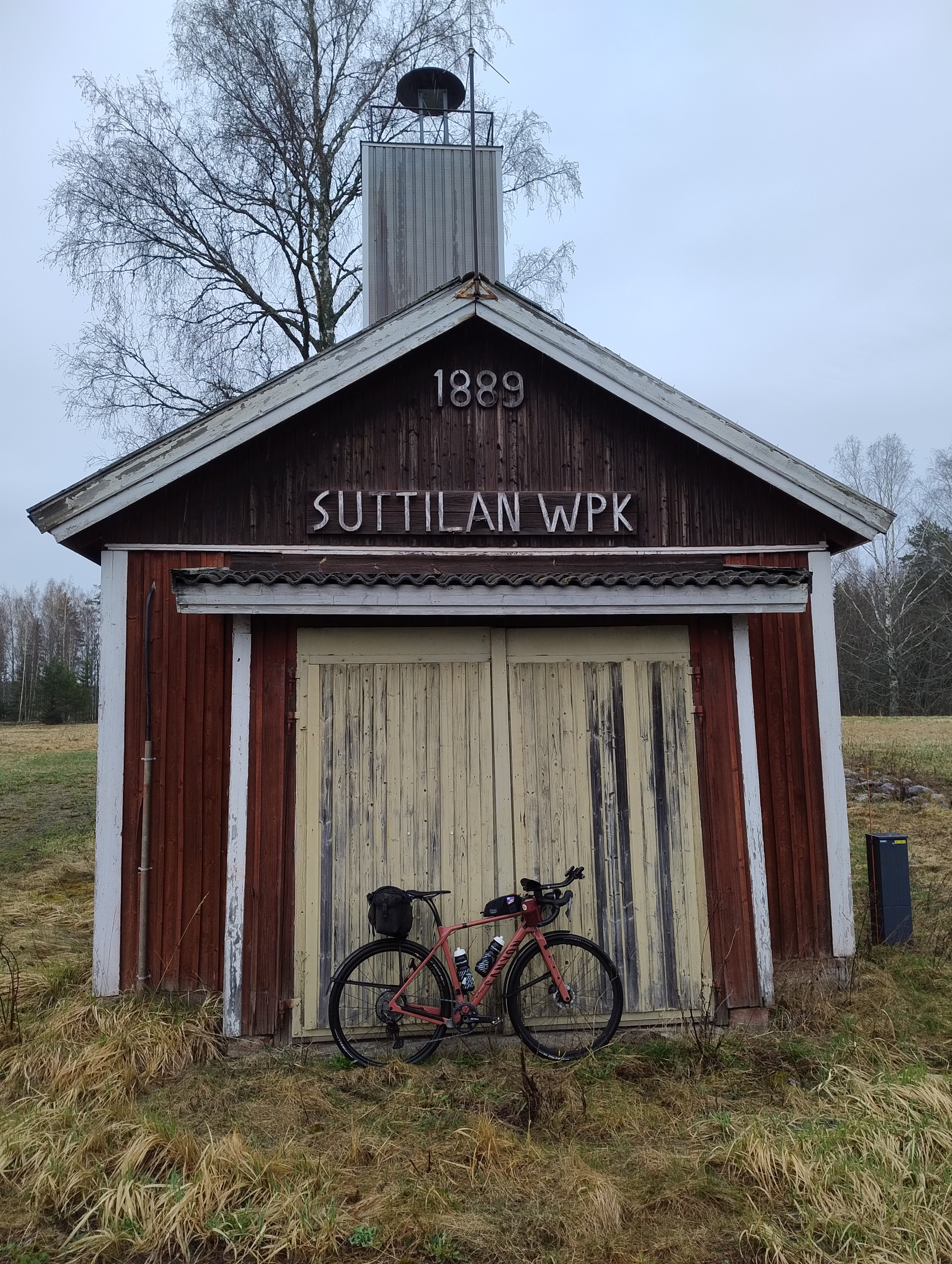 Polkupyörä nojaa punaisen puurakennuksen seinään, yllä kyltti jossa lukee Suttilan WPK ja vuosiluku 1889